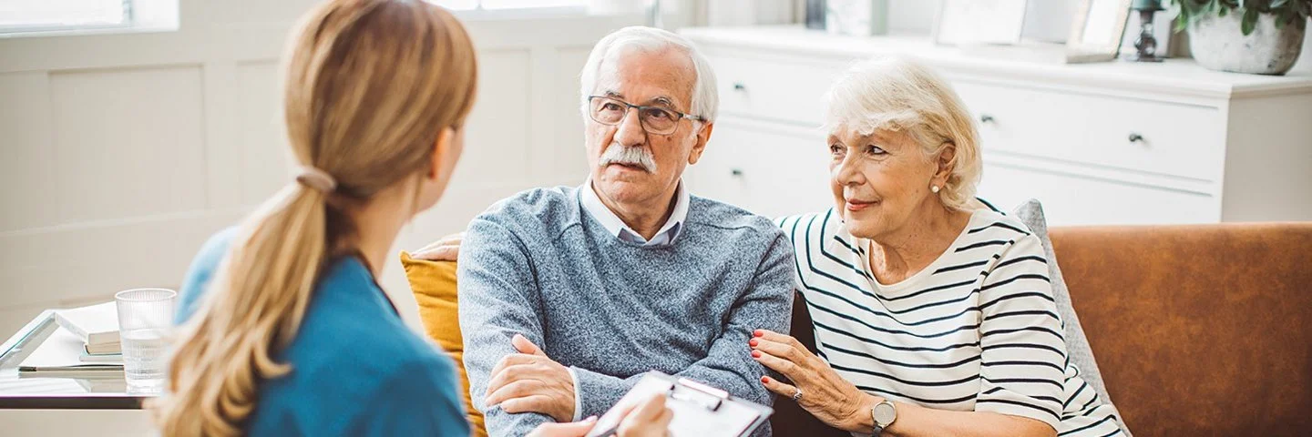Ein älteres Ehepaar sitzt in einem gemütlich eingerichteten Wohnzimmer auf einer braunen Ledercouch. Das Ehepaar unterhält sich mit einer weiblichen Pflegefachkraft, die ihnen gegenübersitzt. Die Pflegefachkraft hält ein Klemmbrett mit einer Liste und einen Stift in der Hand.