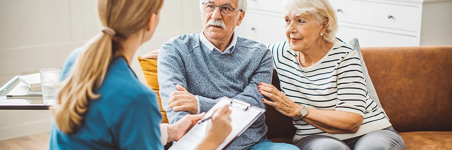 Ein älteres Ehepaar sitzt in einem gemütlich eingerichteten Wohnzimmer auf einer braunen Ledercouch. Das Ehepaar unterhält sich mit einer weiblichen Pflegefachkraft, die ihnen gegenübersitzt. Die Pflegefachkraft hält ein Klemmbrett mit einer Liste und einen Stift in der Hand.