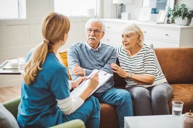 Ein älteres Ehepaar sitzt in einem gemütlich eingerichteten Wohnzimmer auf einer braunen Ledercouch. Das Ehepaar unterhält sich mit einer weiblichen Pflegefachkraft, die ihnen gegenübersitzt. Die Pflegefachkraft hält ein Klemmbrett mit einer Liste und einen Stift in der Hand.