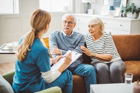 Ein älteres Ehepaar sitzt in einem gemütlich eingerichteten Wohnzimmer auf einer braunen Ledercouch. Das Ehepaar unterhält sich mit einer weiblichen Pflegefachkraft, die ihnen gegenübersitzt. Die Pflegefachkraft hält ein Klemmbrett mit einer Liste und einen Stift in der Hand.