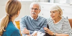 Ein älteres Ehepaar sitzt in einem gemütlich eingerichteten Wohnzimmer auf einer braunen Ledercouch. Das Ehepaar unterhält sich mit einer weiblichen Pflegefachkraft, die ihnen gegenübersitzt. Die Pflegefachkraft hält ein Klemmbrett mit einer Liste und einen Stift in der Hand.