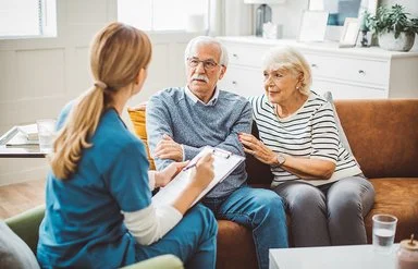 Ein älteres Ehepaar sitzt in einem gemütlich eingerichteten Wohnzimmer auf einer braunen Ledercouch. Das Ehepaar unterhält sich mit einer weiblichen Pflegefachkraft, die ihnen gegenübersitzt. Die Pflegefachkraft hält ein Klemmbrett mit einer Liste und einen Stift in der Hand.