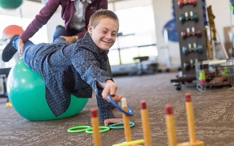 Junge mit Down-Syndrom macht Ergotherapie mithilfe eines Gymnastikballs, um Muskeln aufzubauen.