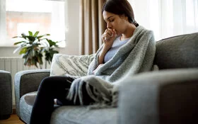 Junge Frau sitzt krank auf der Couch, sie ist in eine Decke gehüllt und hustet in ihre Hand.