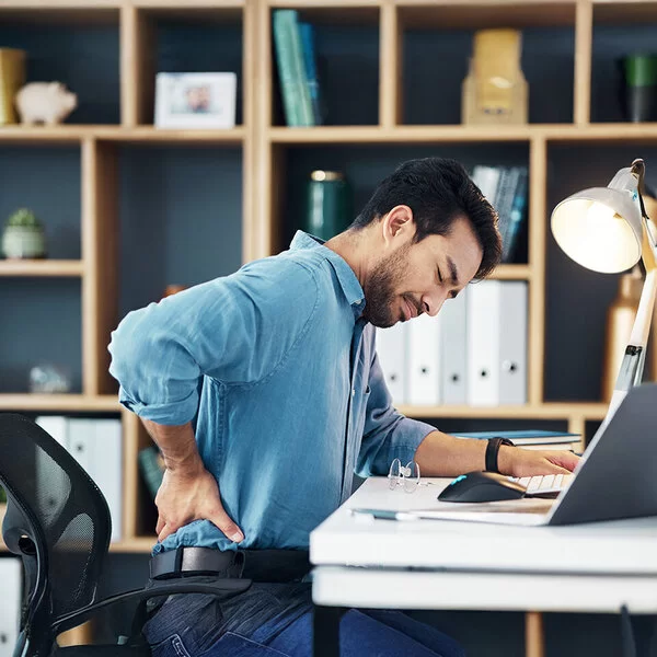 Ein junger Mann sitzt am Schreibtisch in seinem Arbeitszimmer oder Büro. Er hat die Augen geschlossen und fasst sich mit der rechten Hand in die Lendengegend.