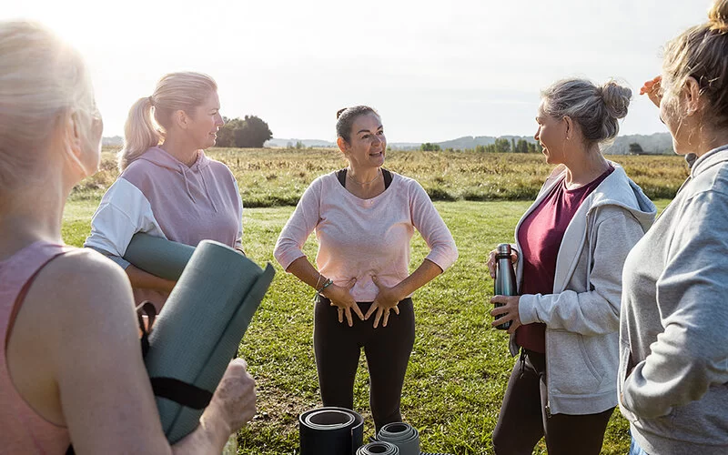 Fünf Frauen um die 50 in sportlicher Kleidung draußen auf einer grünen Wiese. Sie haben Yoga-Matten dabei, eine der Frauen erklärt den anderen eine Übung und deutet mit ihren Händen auf ihre Körpermitte.