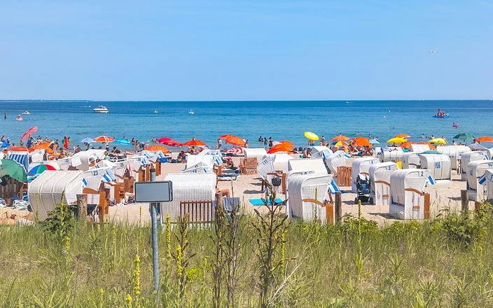 Ein Sandstrand an der Ostsee, der dicht mit Strandkörben und Sonnenschirmen besetzt ist. Die Sonne scheint und das Meer ist ruhig.