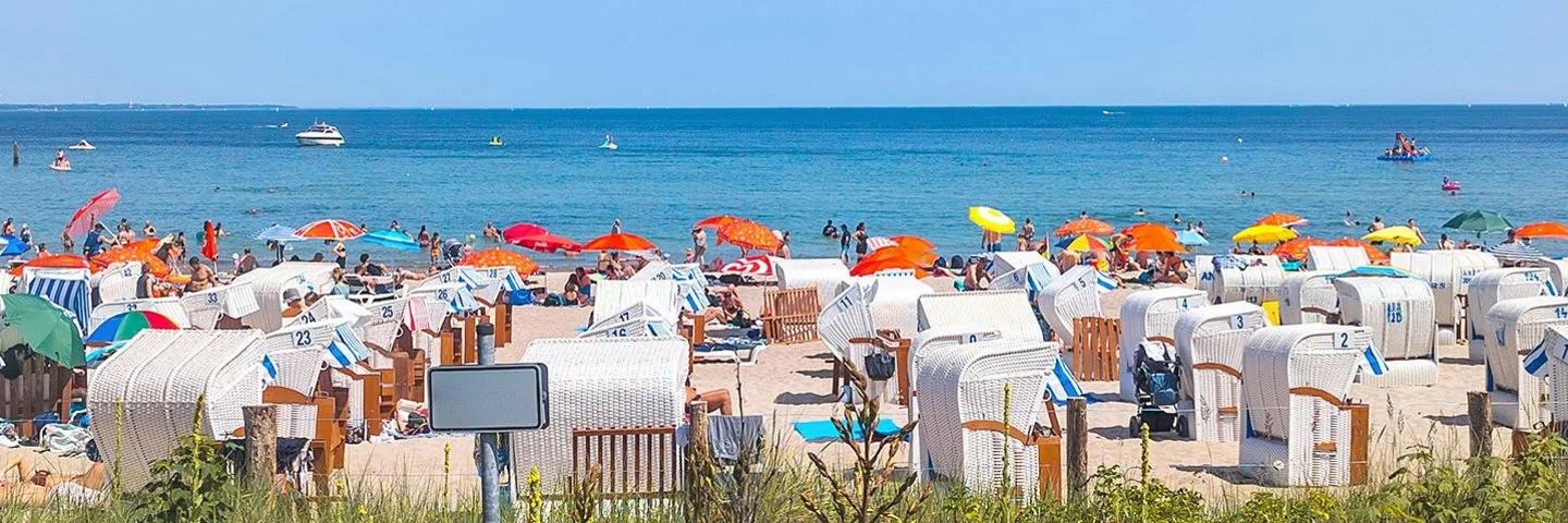 Ein Sandstrand an der Ostsee, der dicht mit Strandkörben und Sonnenschirmen besetzt ist. Die Sonne scheint und das Meer ist ruhig.