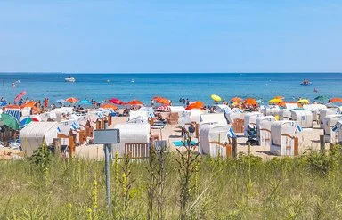 Ein Sandstrand an der Ostsee, der dicht mit Strandkörben und Sonnenschirmen besetzt ist. Die Sonne scheint und das Meer ist ruhig.