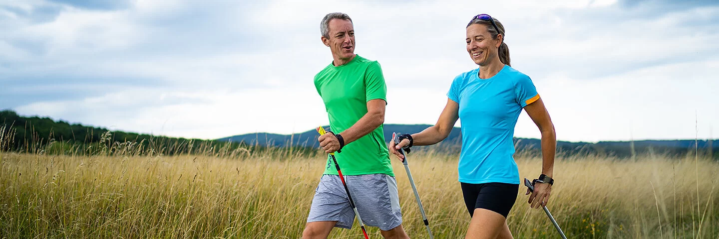 Sport bei MS: Ein Mann und eine Frau mittleren Alters in Shorts und T-Shirts schreiten – ausgerüstet mit Nordic Walking Stöcken – durch offenes Grasland und unterhalten sich dabei. 
