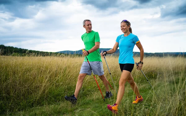 Sport bei MS: Ein Mann und eine Frau mittleren Alters in Shorts und T-Shirts schreiten – ausger�üstet mit Nordic Walking Stöcken – durch offenes Grasland und unterhalten sich dabei. 