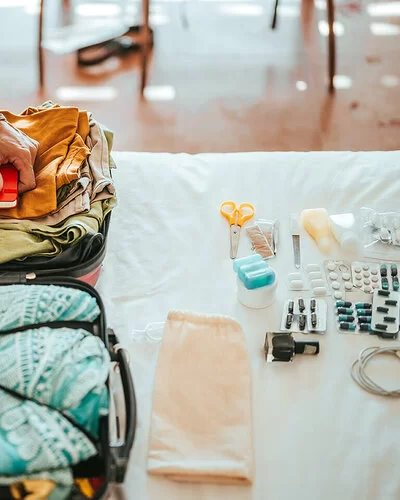 Ein Mann in einem beigefarbenem Hemd und tätowiertem Arm packt Kleidung und alles Notwendige für die Reise in einen Koffer. Vor ihm auf dem Bett liegt sorgfältig aufgereiht der Inhalt seiner Reiseapotheke – von Pflastern bis Tabletten in Blisterpackungen.