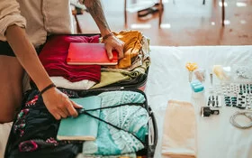 Ein Mann in einem beigefarbenem Hemd und tätowiertem Arm packt Kleidung und alles Notwendige für die Reise in einen Koffer. Vor ihm auf dem Bett liegt sorgfältig aufgereiht der Inhalt seiner Reiseapotheke – von Pflastern bis Tabletten in Blisterpackungen.