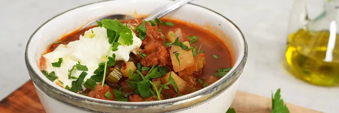 Veganer Gemüseeintopf nach einem Rezept von Felicitas Then, serviert in einer hellen Schüssel mit einem Klecks veganem Crème fraîche-Topping. Rechts im Hintergrund ein Ölkännchen, Kräuter und ein Holzbrett.