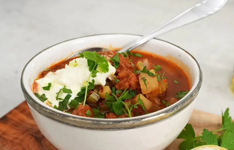 Veganer Gemüseeintopf nach einem Rezept von Felicitas Then, serviert in einer hellen Schüssel mit einem Klecks veganem Crème fraîche-Topping. Rechts im Hintergrund ein Ölkännchen, Kräuter und ein Holzbrett.