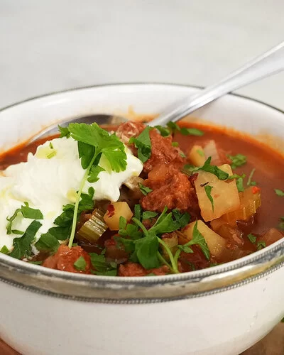 Veganer Gemüseeintopf nach einem Rezept von Felicitas Then, serviert in einer hellen Schüssel mit einem Klecks veganem Crème fraîche-Topping. Rechts im Hintergrund ein Ölkännchen, Kräuter und ein Holzbrett.
