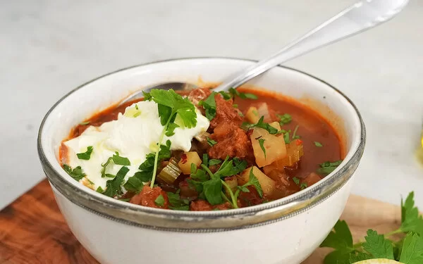 Veganer Gemüseeintopf nach einem Rezept von Felicitas Then, serviert in einer hellen Schüssel mit einem Klecks veganem Crème fraîche-Topping. Rechts im Hintergrund ein Ölkännchen, Kräuter und ein Holzbrett.