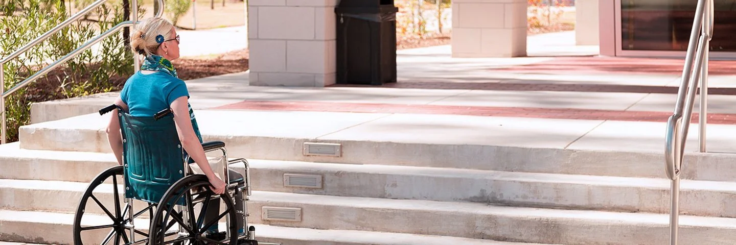 Eine mittelalte Frau im Rollstuhl steht bei sonnigem Wetter draußen vor einigen Stufen, die zu einem Gebäude führen. Es ist keine Rampe in Sicht.