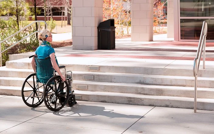 Eine mittelalte Frau im Rollstuhl steht bei sonnigem Wetter draußen vor einigen Stufen, die zu einem Gebäude führen. Es ist keine Rampe in Sicht.