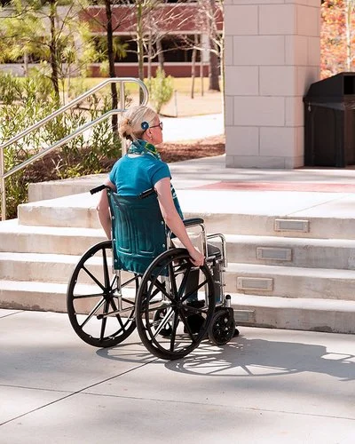 Eine mittelalte Frau im Rollstuhl steht bei sonnigem Wetter draußen vor einigen Stufen, die zu einem Gebäude führen. Es ist keine Rampe in Sicht.
