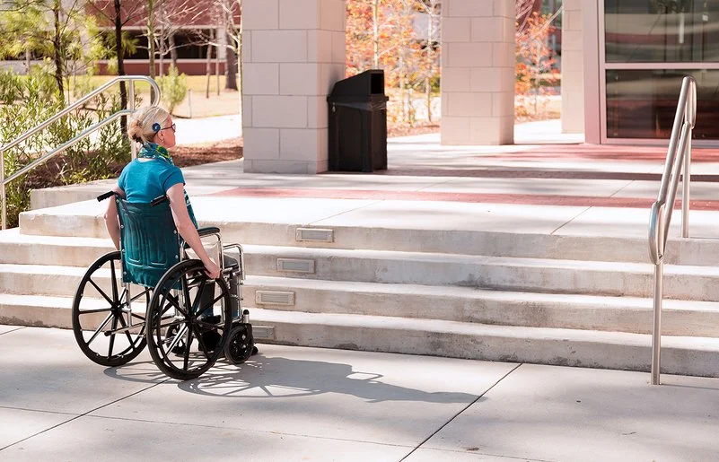 Eine mittelalte Frau im Rollstuhl steht bei sonnigem Wetter draußen vor einigen Stufen, die zu einem Gebäude führen. Es ist keine Rampe in Sicht.