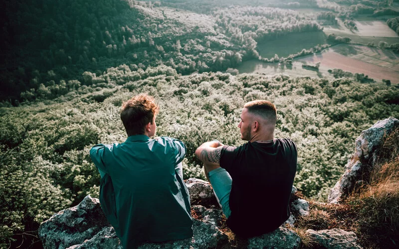 Zwei junge Männer sitzen auf einem Felsvorsprung und schauen über Wälder und Felder.