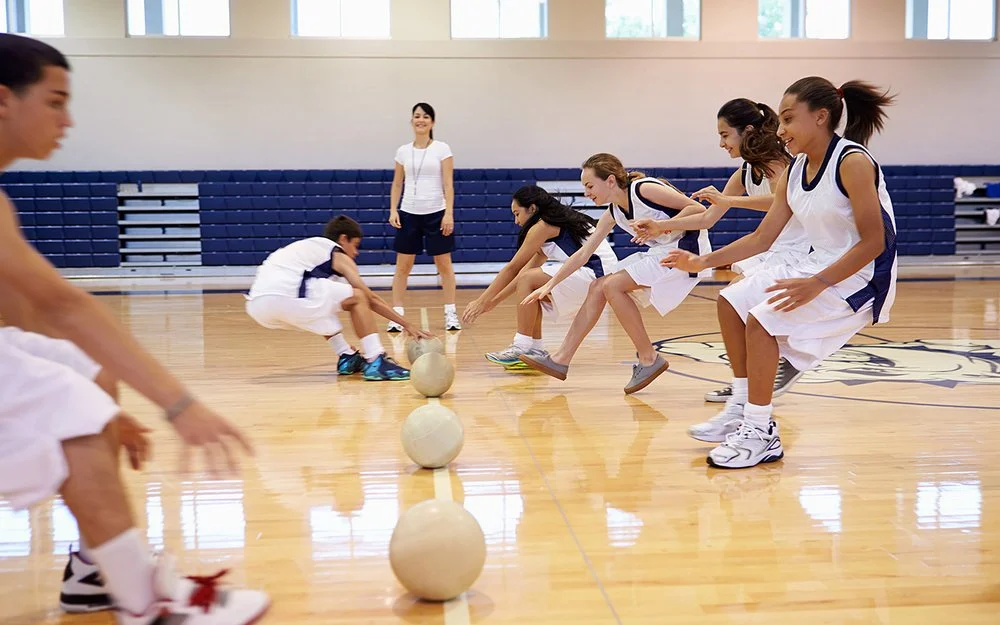 Zwei Teams bei einem Dodgebal-Spiel in einer Turnhalle. Beide Seiten laufen auf die in der Mitte liegenden Bälle zu und greifen danach.