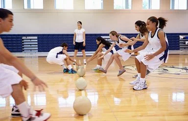 Zwei Teams bei einem Dodgebal-Spiel in einer Turnhalle. Beide Seiten laufen auf die in der Mitte liegenden Bälle zu und greifen danach.