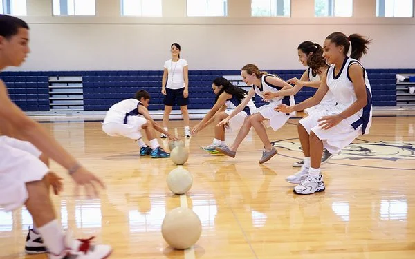 Zwei Teams bei einem Dodgebal-Spiel in einer Turnhalle. Beide Seiten laufen auf die in der Mitte liegenden Bälle zu und greifen danach.