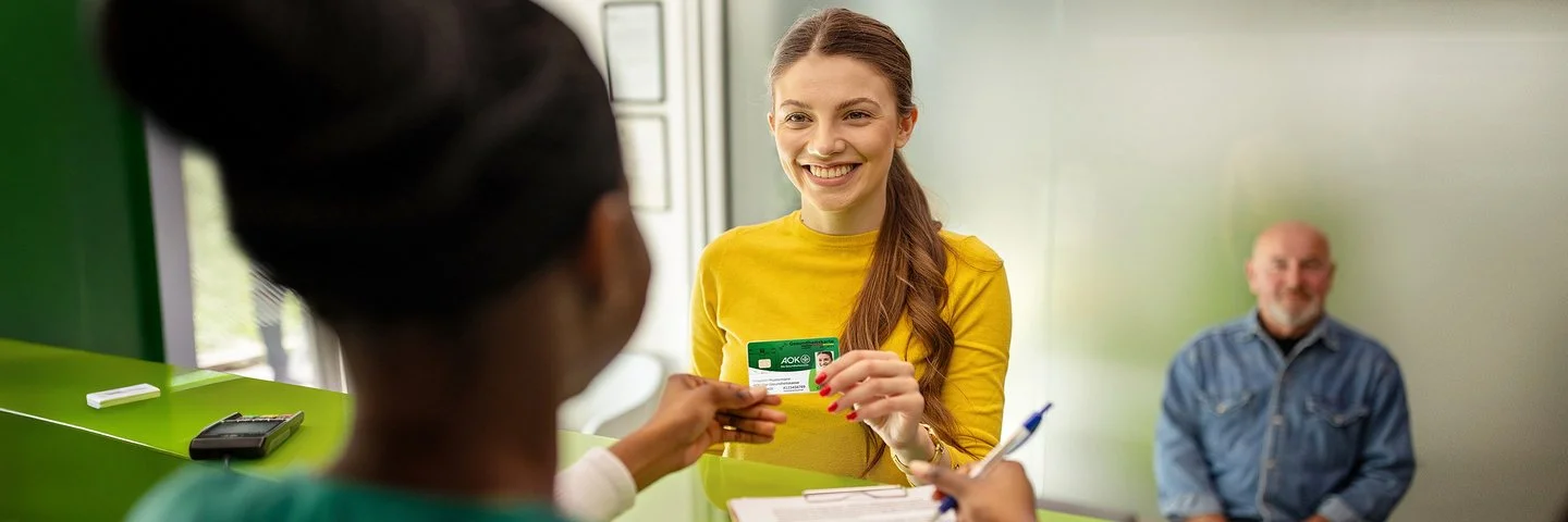Eine Frau mit langen, braunen Haaren trägt ein gelbes Oberteil und hält eine elektronische Gesundheitskarte der AOK in der Hand. Sie steht an einem Tresen und reicht die Karte einer Person mit zusammengebundenen Haaren, die auf der anderen Seite des Tresens sitzt. Im Hintergrund sitzt eine weitere Person auf einem Stuhl vor einer grünen Wand. Auf dem Tresen liegt ein Kartenlesegerät.
