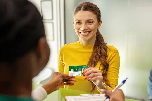 Eine Frau mit langen, braunen Haaren trägt ein gelbes Oberteil und hält eine elektronische Gesundheitskarte der AOK in der Hand. Sie steht an einem Tresen und reicht die Karte einer Person mit zusammengebundenen Haaren, die auf der anderen Seite des Tresens sitzt. Im Hintergrund sitzt eine weitere Person auf einem Stuhl vor einer grünen Wand. Auf dem Tresen liegt ein Kartenlesegerät.