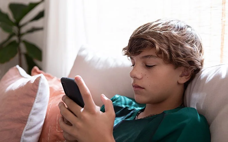 Ein Teenager in einem grünen T-Shirt sitzt auf dem Sofa und schaut ins Smartphone.