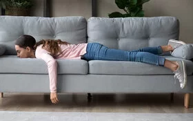 Eine junge Frau mit Langeweile liegt auf dem Bauch auf dem Sofa und lässt die Arme herunterhängen.