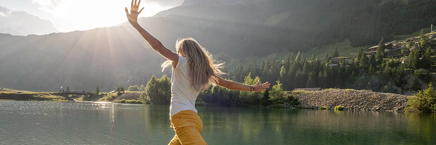 Eine junge Frau mit langen blonden Haaren, weißem T-Shirt und gelber langer Hose springt auf einen Stein in einem See. Sie breitet die Arme zur Seie aus und streckt ein Bein im Sprung nach hinten. Das Wasser des Sees ist klar, man kann den Boden sehen. Im Hintergrund sind grüne Bäume und Berge zu sehen. 