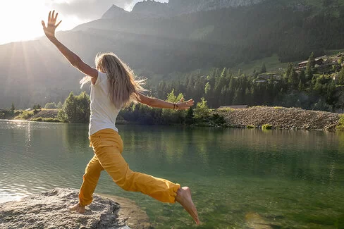 Eine junge Frau mit langen blonden Haaren, weißem T-Shirt und gelber langer Hose springt auf einen Stein in einem See. Sie breitet die Arme zur Seie aus und streckt ein Bein im Sprung nach hinten. Das Wasser des Sees ist klar, man kann den Boden sehen. Im Hintergrund sind grüne Bäume und Berge zu sehen. 
