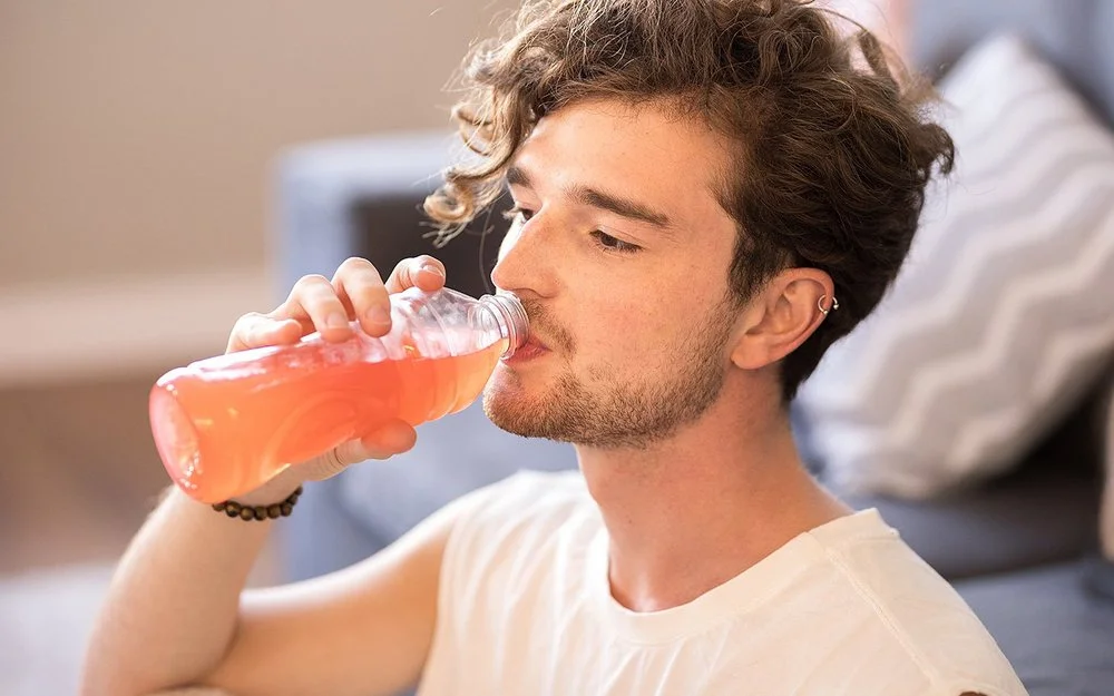 Junger Mann mit lockigem Haar sitzt im Wohnzimmer und trinkt eine Flasche dezent rosa gefärbtes Vitaminwasser.