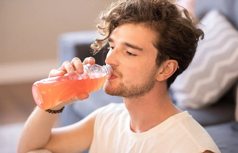 Junger Mann mit lockigem Haar sitzt im Wohnzimmer und trinkt eine Flasche dezent rosa gefärbtes Vitaminwasser.