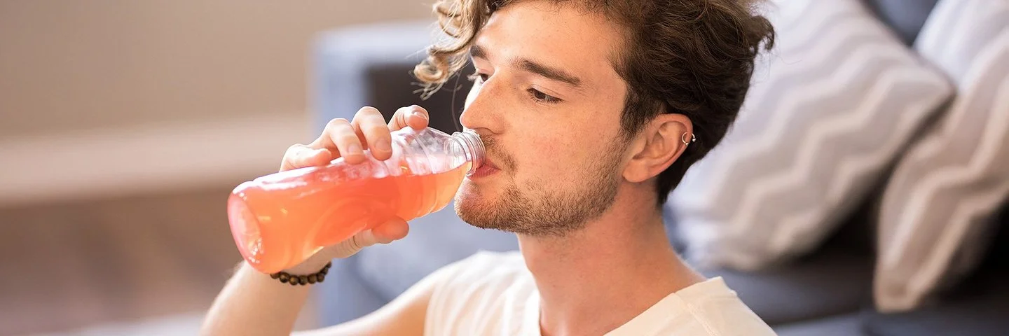 Junger Mann mit lockigem Haar sitzt im Wohnzimmer und trinkt eine Flasche dezent rosa gefärbtes Vitaminwasser.