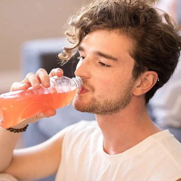 Junger Mann mit lockigem Haar sitzt im Wohnzimmer und trinkt eine Flasche dezent rosa gefärbtes Vitaminwasser.
