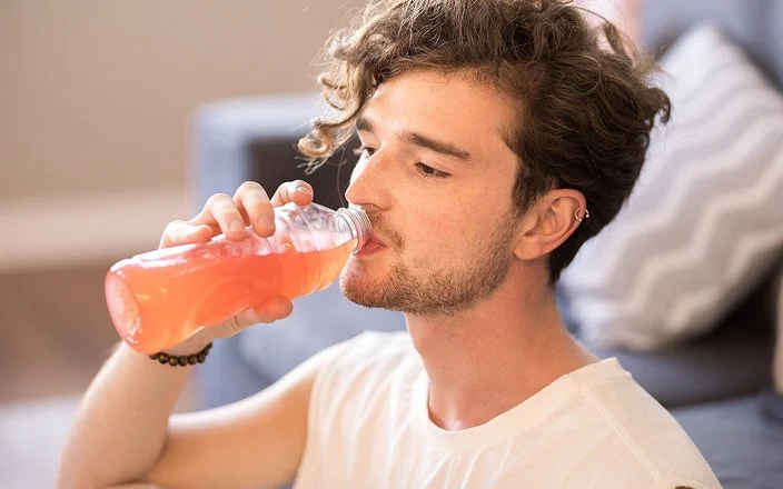 Junger Mann mit lockigem Haar sitzt im Wohnzimmer und trinkt eine Flasche dezent rosa gefärbtes Vitaminwasser.