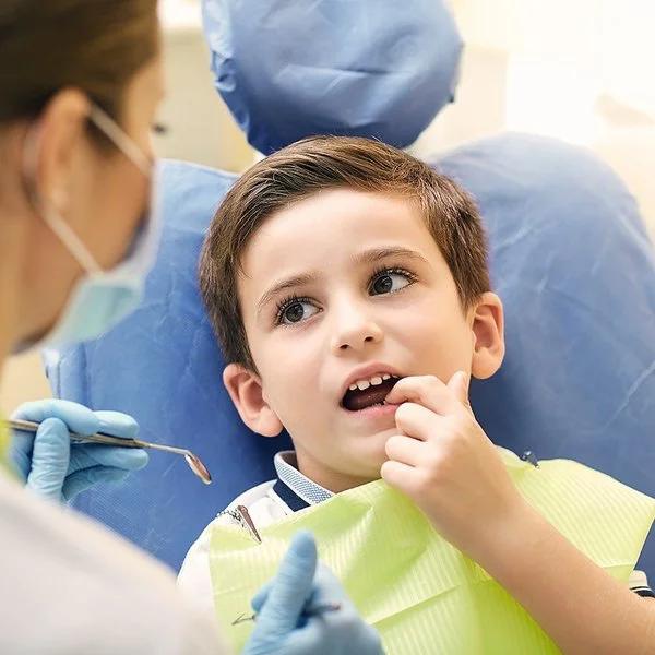 Ein kleiner Junge sitzt mit geöffnetem Mund auf dem Behandlungsstuhl einer Zahnarztpraxis. Er zeigt mit dem Finger auf einen Zahn und schaut dabei zu einer Zahnärztin, die sich in Rückenansicht zeigt.