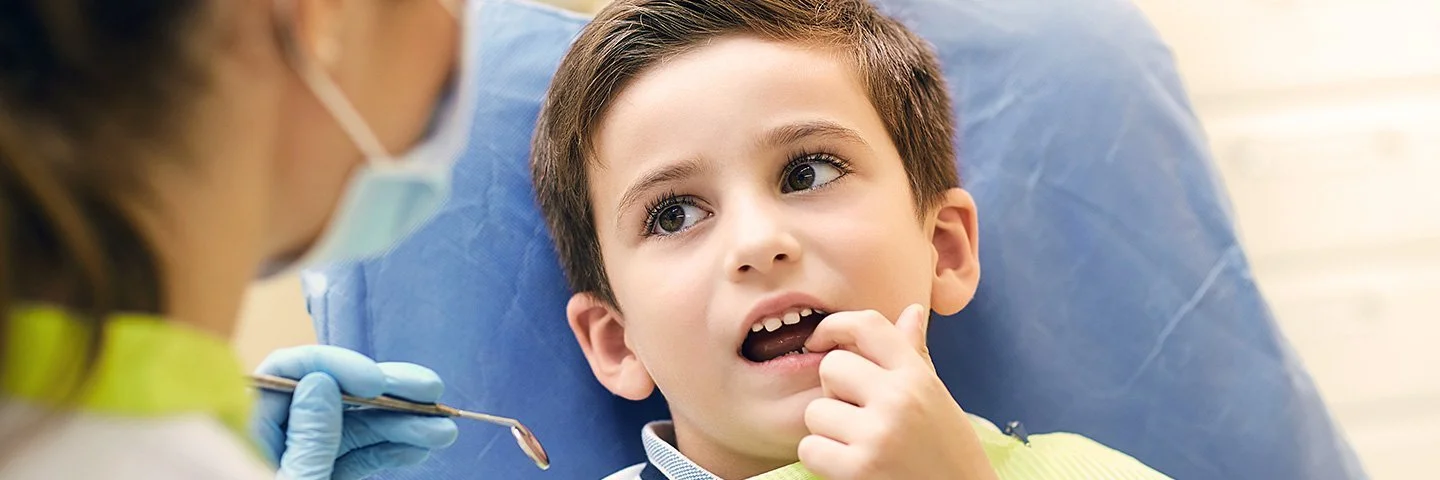 Ein kleiner Junge sitzt mit geöffnetem Mund auf dem Behandlungsstuhl einer Zahnarztpraxis. Er zeigt mit dem Finger auf einen Zahn und schaut dabei zu einer Zahnärztin, die sich in Rückenansicht zeigt.