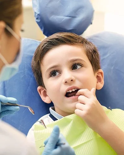 Ein kleiner Junge sitzt mit geöffnetem Mund auf dem Behandlungsstuhl einer Zahnarztpraxis. Er zeigt mit dem Finger auf einen Zahn und schaut dabei zu einer Zahnärztin, die sich in Rückenansicht zeigt.
