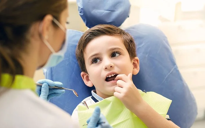 Ein kleiner Junge sitzt mit geöffnetem Mund auf dem Behandlungsstuhl einer Zahnarztpraxis. Er zeigt mit dem Finger auf einen Zahn und schaut dabei zu einer Zahnärztin, die sich in Rückenansicht zeigt.