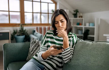 Eine junge Frau sitzt auf ihrem Sofa und schaut besorgt auf ihr Handy.