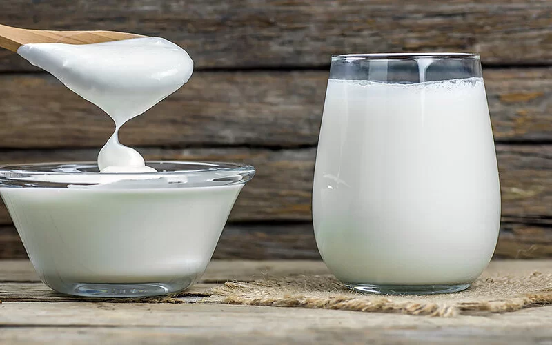 Ein Glas Ayran steht auf einem Tisch. Daneben eine Schüssel mit Joghurt. Mit einem Holzlöffel wird noch mehr hineingegeben.