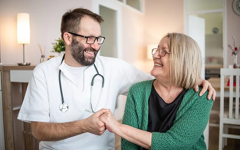Ein Mann in weißer medizinischer Arbeitskleidung mit einem Stethoskop um den Hals sitzt neben einer Frau und hält ihre Hand.