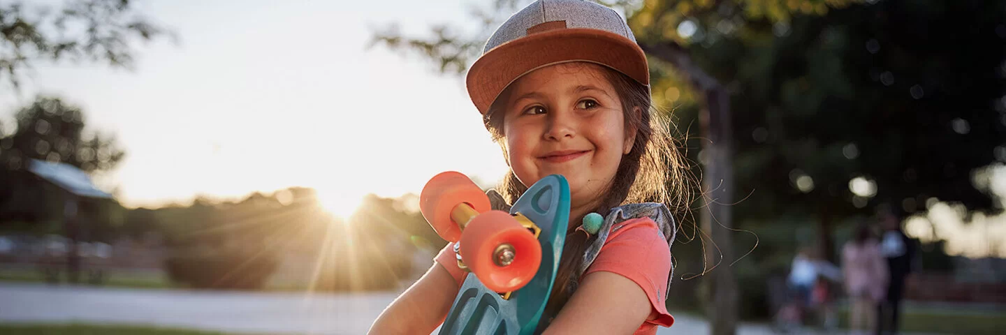 Ein etwa fünfjähriges Mädchen mit Schirmmütze und Latzhose steht auf einer Wiese in einem Park, im Hintergrund ist ein asphaltierter Weg zu erkennen. Das Mädchen hält ein Skateboard mit beiden Armen vor dem Oberkörper und blick zuversichtlich nach vorn.