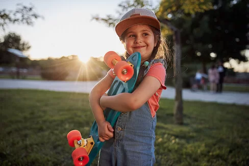 Ein etwa fünfjähriges Mädchen mit Schirmmütze und Latzhose steht auf einer Wiese in einem Park, im Hintergrund ist ein asphaltierter Weg zu erkennen. Das Mädchen hält ein Skateboard mit beiden Armen vor dem Oberkörper und blick zuversichtlich nach vorn.