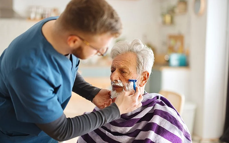 Ein junger Mann in einem Pflegekittel rasiert einem älteren Mann mit einer Rasierklinge den Bart.